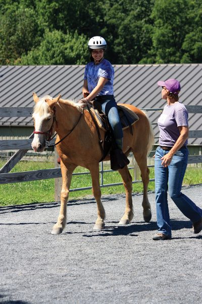 A trainer walks alongside a rider at summer horse camp