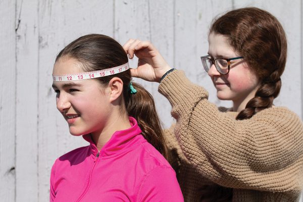 A young girl measures her friends head with a measuring tape to help her find the right fit for a riding helmet