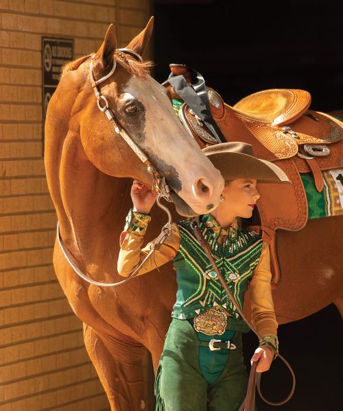 A young equestrian in western show attire holds the reins to a sorrel horse in western tack