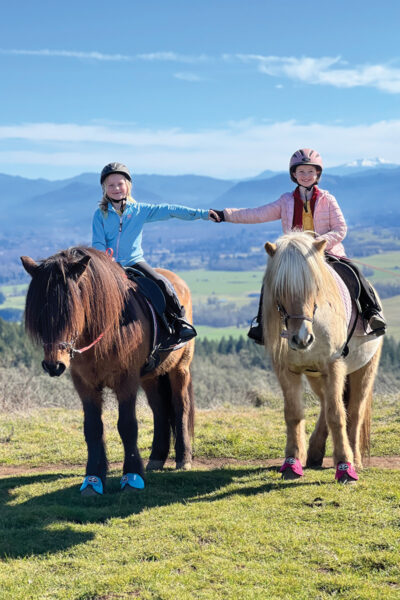 Two young riders hold hands while aboard their ponies in the mountains