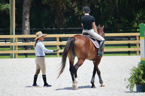 An equestrian riding a lesson horse and receiving instruction from her trainer.