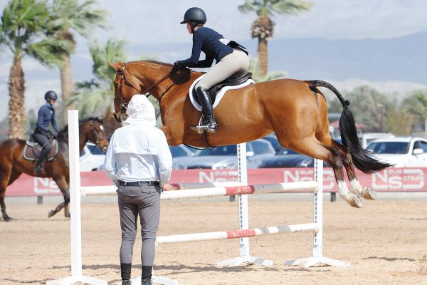 A trainer watches as an equestrian jumps her horse